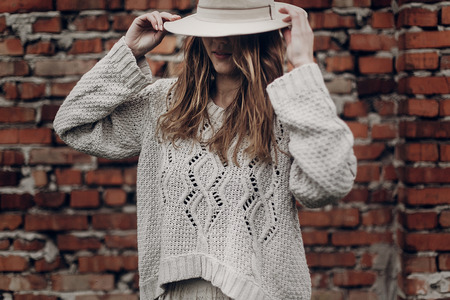 Stylish brunette woman in white hat and boho white sweater posing near red brick wallの写真素材