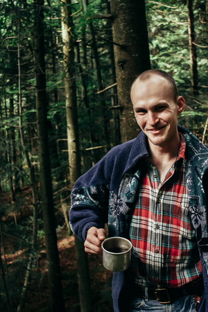 stylish hipster traveler drinking from cup in sunny forest in the mountains