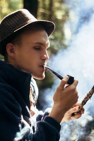 stylish hipster traveler smoking tobacco pipe in sunny forest in the mountainsの写真素材