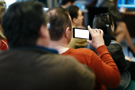 stylish man holding phone with empty white screen in audience and recording, sitting at meeting, business marketing lectureの写真素材