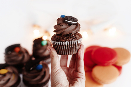 stylish chocolate cupcake in hand  with cookie and candy on top a on white background with macaroons and christmas lights. space for text. catering for celebrations and holidays conceptの写真素材