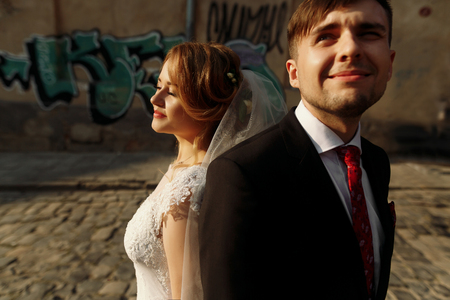 Beautiful smiling bride in vintage white wedding dress leaning against handsome smiling groom outdoors in street at sunset, family portrait closeupの写真素材
