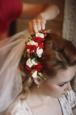 Beautiful bride putting on floral head wreath, stylist doing hairstyle for blonde woman in stylish robe, morning wedding preparation face closeupの写真素材