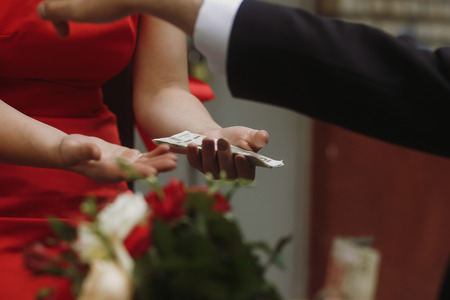 Handsome groom in elegant black suit and wedding bouquet paying for the bride to bridesmaid, slavic wedding tradition, money closeupの写真素材
