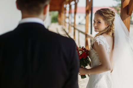 Emotional blonde bride smiling & looking at groom, elegant bride in white wedding dress posing outdoors with bouquetの写真素材