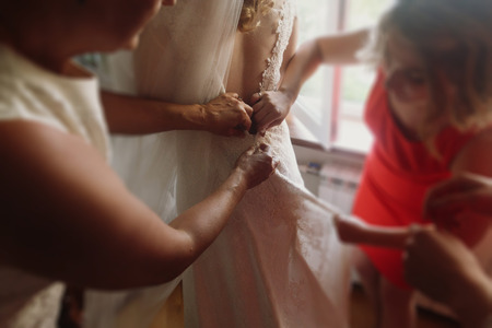 Beautiful bridesmaids help stylish bride put on wedding dress, buttoning lace dress hands closeup, morning preparation for wedding ceremonyの写真素材