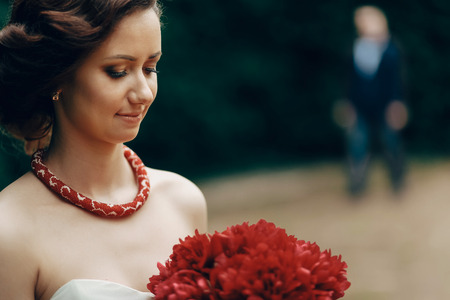 Portrait of gorgeous brunette bride in elegant white dress with red flowers bouquet posing in summer park, handsome groom in the background, newlywed woman momentの写真素材