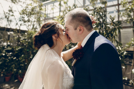 Happy groom kissing with beautiful brunette bride in botanical garden during summer walk, newlywed couple portrait outdoorsの写真素材