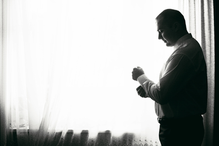 Handsome young man preparing for business meeting, putting on shirt, happy groom posing near window in hotel room before the wedding, businessman conceptの写真素材