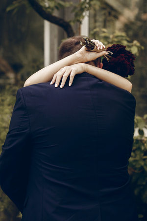 Cheerful newlywed couple hugging outdoors in summer park near trees, handsome groom embracing beautiful bride with bouquetの写真素材