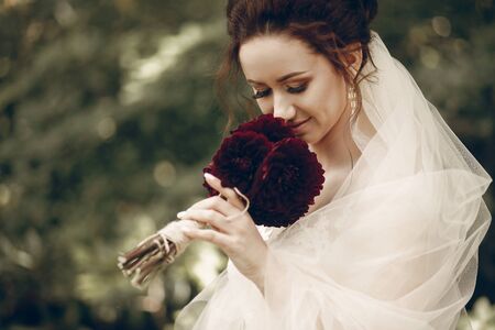 Portrait of beautiful brunette bride in vintage white wedding dress holding fresh red flowers bouquet, posing in the park, newlywed bride conceptの写真素材