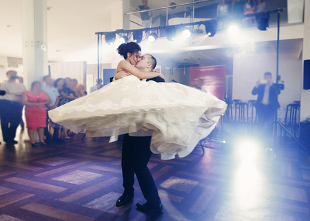 Romantic newlywed couple first dance at wedding reception with guests in the background, groom holding bride in the air and swirling at dance hallの写真素材