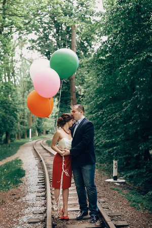 Beautiful portrait of fun newlywed couple with balloons, handsome groom kissing bride on forehead near old railway in a park, travel conceptの写真素材