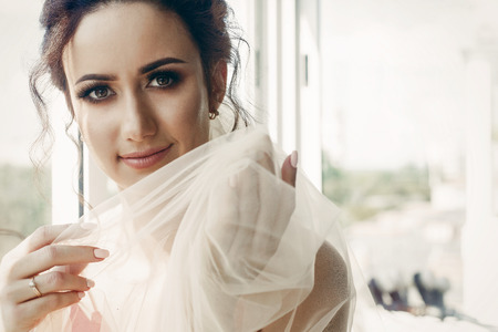 Beautiful emotional bride in sexy red robe with white wedding veil portrait near window, morning before the wedding, gorgeous brunette woman face close-upの写真素材