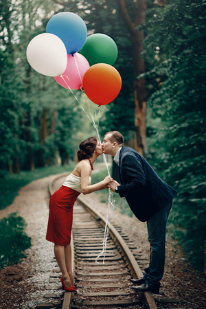 Romantic moment - happy couple kissing on railroad and holding colorful ballons, newlywed bride and groom holding hands and kissing outdoors, honeymoon conceptの写真素材