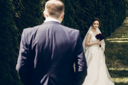 Beautiful bride with red flowers bouquet looking at stylish groom walking towards her, happy newlywed couple posing outdoors near row of treesの写真素材