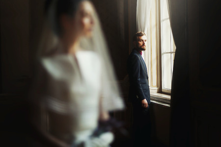 elegant stylish handsome groom looking at gorgeous bride, standing at window light. unusual luxury wedding couple in retro styleの写真素材