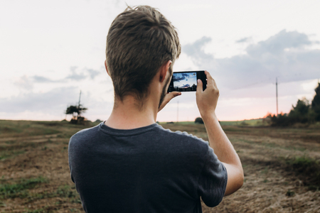 stylish hipster traveler holding smart phone taking photo of beautiful sunset landscape in summer field. exploring and discovering. space for textの写真素材
