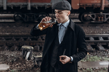 stylish gangster man drinking. posing on background of railway. england in 1920s theme. fashionable brutal confident guy. atmospheric  moments. space for textの写真素材