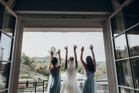 luxury gorgeous bride and her bridesmaids having fun in hotel room, at window, wedding morning. hands up in the air and cheering. back view. space for text. joyful momentの写真素材