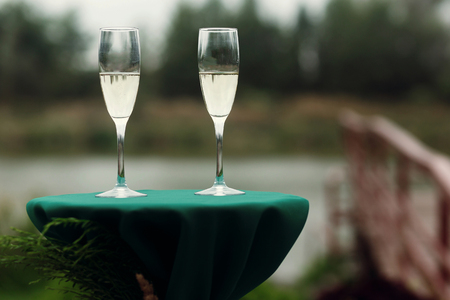 Two luxury champagne glasses at green table at wedding aisle, outdoors near wooden bridge and lake, two alcohol drinks closeup, wedding ceremony decorationの写真素材