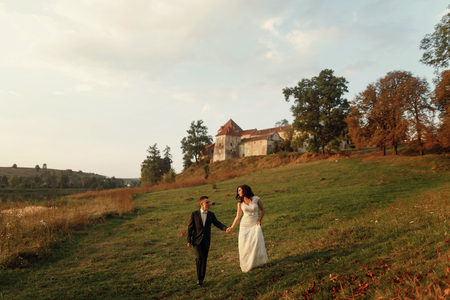 gorgeous bride and stylish groom walking holding hands in soft evening light at sunset near castle. sensual romantic moment of beautiful wedding couple outdoorsの写真素材