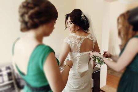 Stylish beautiful bridesmaids helping gorgeous brunette bride in white dress get ready for wedding, morning preparations, woman putting on dressの写真素材