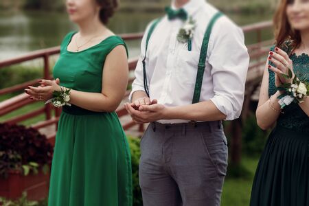 Happy beautiful guests at wedding ceremony, stylish groomsman and gorgeous bridesmaids in green dress with white boutonnieres applaud at outdoor ceremony, hands closeupの写真素材