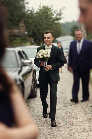 Handsome happy brunette groom in stylish black suit with white roses wedding bouquet walking to office for ceremony with guests and groomsmenの写真素材