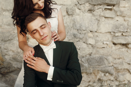 stylish luxury bride and groom embracing gently together near old castle wall at sunset. sensual moment of beautiful wedding couple outdoorsの写真素材