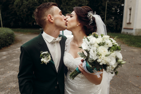 Gorgeous brunette bride in lace white wedding dress kissing handsome happy groom in stylish suit, newlyweds walking in park portrait, face closeupの写真素材