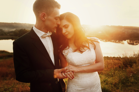 gorgeous bride and stylish groom gently hugging in the evening warm sunlight. sensual romantic moment of luxury wedding couple in amazing sunsetの写真素材