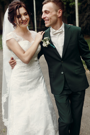 Happy newlywed couple walking down summer park road, cheerful handsome groom in stylish suit hugging happy, smiling bride in elegant white wedding dressの写真素材