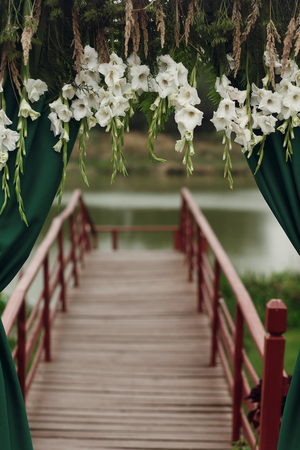 Beautiful stylish wedding aisle pathway with white floral garland hanging from arc with green fashionable curtain near wooden bridge at lake, flowers closeupの写真素材