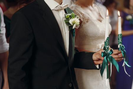 gorgeous wedding couple holding candles at official ceremony in old church. traditional moment of marriage of bride and stylish groomの写真素材