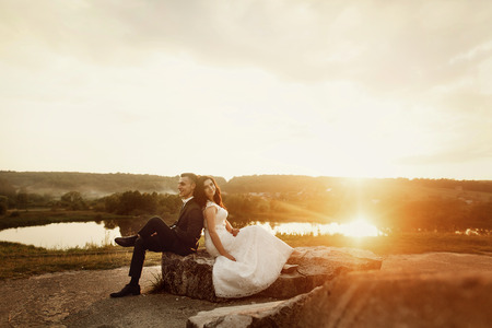 Romantic couple of newlyweds resting on stone bench at sunset field near lake landscape, happy bride leaning against smiling groom outdoorsの写真素材