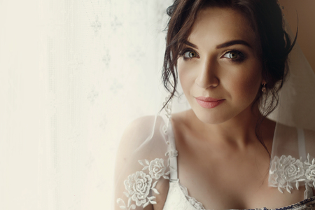 Portrait of beautiful brunette bride in luxury white wedding dress posing near window, morning wedding preparation, face closeupの写真素材
