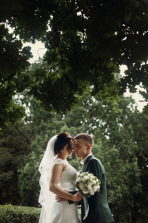 Romantic newlywed couple hugging under tree in park, handsome sensual groom with beautiful brunette bride in vintage white dress with white roses bouquetの写真素材