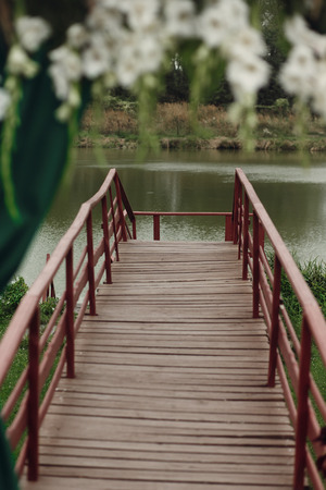 Beautiful stylish wedding aisle pathway with white flowers garland hanging from arc with green fashionable curtain near wooden bridge at lakeの写真素材