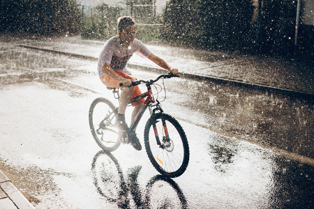 young boy riding bicycle in rainy street in sunshine, summer moments. space for text. atmospheric moment. cycling and activity. stylish hipster having fun under rainの写真素材