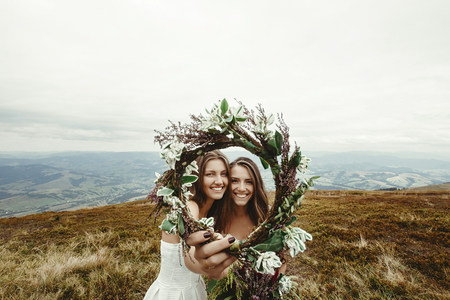 stylish bridesmaid and gorgeous bride laughing and looking through wreath, boho wedding, luxury ceremony at mountainsの写真素材