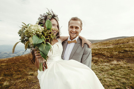 stylish groom carrying happy bride and laughing, boho wedding couple, luxury ceremony at mountains with amazing viewの写真素材
