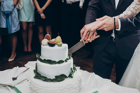 happy wedding couple cutting together wedding cake. rustic cake with greenery pear and figs and macaroons at luxury wedding reception in restaurant. expensive catering. space for textの写真素材