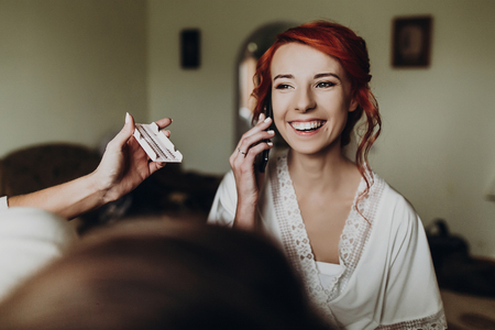 beautiful stylish bride with red hair talking on phone and smiling, wearing robe,  getting make up done by professional, morning preparation. wedding getting ready, space for textの写真素材