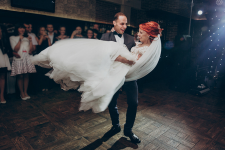 stylish groom lifting happy bride and  dancing at wedding reception in light. gorgeous wedding couple performing first dance in restaurant. newlyweds, emotional moment. space for textの写真素材