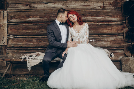 rustic wedding couple sitting on bench at wooden house in evening. space for text. stylish groom and happy bride dreaming, newlyweds embracing, sensual romantic momentの写真素材