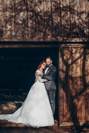 rustic wedding couple posing and hugging in sunlight at background of wooden wall in country barn. wedding concept, space for text. happy stylish bride and groom embracing, bohemian newlywedsの写真素材
