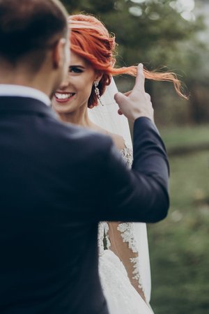 stylish groom embracing his happy gorgeous bride and touching her hair gently in park. romantic sensual moment, space for text. luxury wedding newlyweds coupleの写真素材