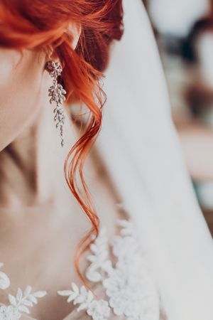 stylish bride detail, luxury earring and red hair curl, beautiful wedding dress. rustic wedding morning preparation. bridal getting ready. emotional moment. space for textの写真素材