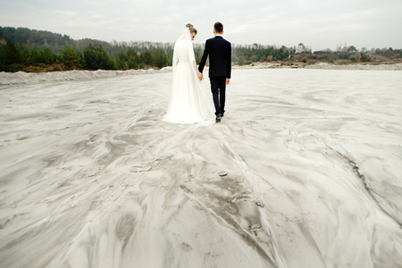 gorgeous bride and groom walking holding hands and looking ar each other at sandy beach lake, true emotionsの写真素材